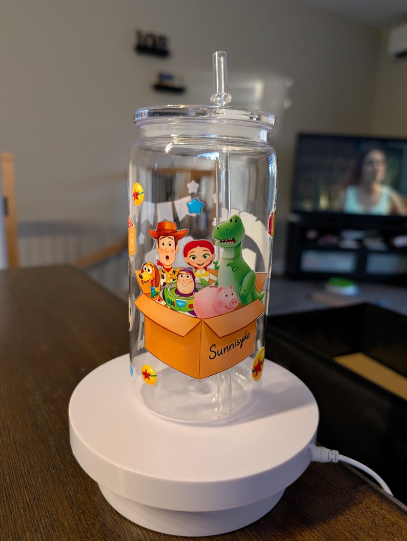 May include: A clear glass tumbler with a lid and straw featuring a colorful graphic of cartoon characters including a cowboy, space ranger, cowgirl, green dinosaur, and pink pig inside a cardboard box labeled Sunnyside with small star accents.
