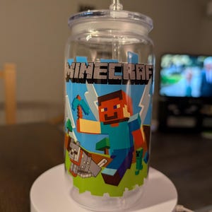 以下が含まれることがあります： Minecraftのデザインが施された、蓋とストロー付きの透明なプラスチックタンブラー。デザインには、青とグレーの「Minecraft」の文字、ピクセル化されたキャラクター、緑と青の背景が含まれています。タンブラーは白いスタンドの上にあります。