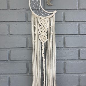 May include: Cream-colored macrame dream catcher with a crescent moon design. The top features a crescent moon shape with a webbed interior and a small, hanging moon charm. Long, knotted fringe hangs below, accented with wooden beads.