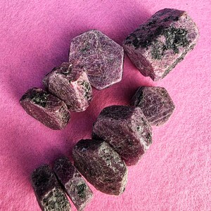 May include: A collection of rough-cut ruby gemstones in varying sizes. The stones display a deep reddish-purple hue with dark inclusions, set against a bright pink background. The gemstones have a natural, unpolished appearance.