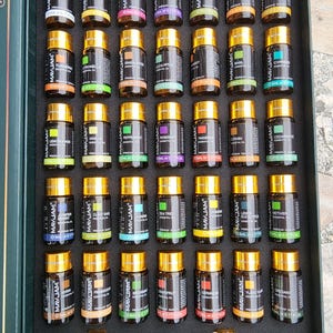 May include: A set of essential oil bottles in a dark green box. The small, brown glass bottles have gold caps and labels with various names. Two droppers are included. The oils are arranged in rows, showcasing a variety of scents.