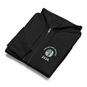 Può includere: Felpa con cappuccio nera con cerniera, cappuccio e cerniera bianca. La felpa con cappuccio presenta una grafica circolare con il testo "TWO NATIONS ONE GOAL" e l'anno "2026". La grafica include le bandiere americana e messicana.