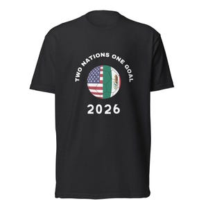 Può includere: T-shirt nera con un design a pallone da calcio che incorpora le bandiere di Stati Uniti e Messico. Il testo "TWO NATIONS ONE GOAL" circonda il pallone, con "2026" sotto. Una maglietta casual per i tifosi di calcio.