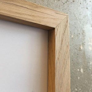 May include: Close-up of the corner of a natural wood picture frame. The frame is made of light brown wood with a visible grain pattern.