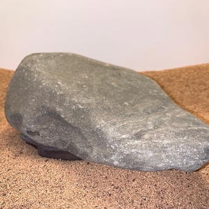 May include: A large, gray, irregularly shaped rock resting on a bed of brown sand.