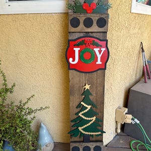 May include: A wooden Christmas sign featuring a red and green JOY sign, a green Christmas tree with gold garland, and a holly sprig with red berries. The sign has three black circles for hanging decorations.