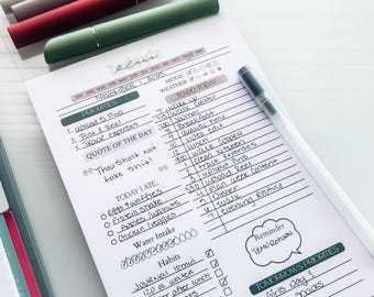 May include: A daily planner with a to-do list, priorities, and a quote. Several pens in green, red, and brown are arranged above the planner. A clear pen is on the right side of the planner.