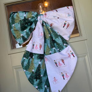 May include: A large fabric bow featuring two distinct patterns: one with green pine trees and white stars on a teal background, and another with nutcracker figures and ballerinas in pink dresses on a white background, hanging from a clear hook.