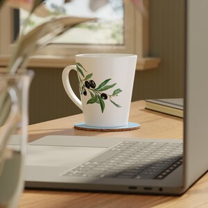 May include: White ceramic mug with a green olive branch design. The mug is sitting on a blue coaster on a wooden table next to a laptop.