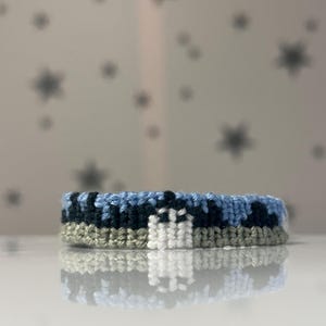 Könnte beinhalten: Ein gewebtes Freundschaftsarmband mit einem horizontalen Muster aus hellblauem Himmel, einer dunkelblauen Bergkette und einem hellgrauen Boden. Eine kleine weiße rechteckige Form befindet sich mittig im dunkelblauen Gebirgsabschnitt.