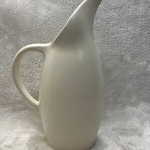 May include: A white ceramic pitcher with a curved spout and a handle. The pitcher has a smooth, matte finish.