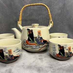 May include: A white ceramic tea set with a bamboo handle on the teapot. The set features a blue and red design of a warrior on horseback. The set includes a teapot and four teacups.