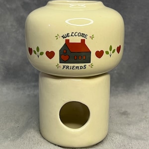 May include: A white ceramic candle holder with a house design and the words "Welcome Friends". The holder has a red roof, red hearts, and green leaves.