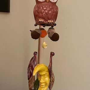 May include: A brown ceramic owl with yellow eyes hangs from a wooden dowel. Below the owl are brown, orange, and yellow ceramic acorns.  The owl is part of a wind chime with brown, yellow, and green ceramic leaves and a yellow ceramic crescent moon with a face.