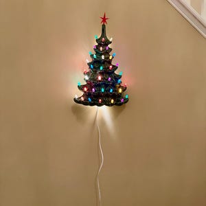 Könnte beinhalten: Dunkelgrüner Keramik-Weihnachtsbaum zur Wandmontage mit bunten Lichtern und einem roten Stern. Er ist an einer beigen Wand befestigt und hat ein weißes Stromkabel. Eine festliche Weihnachtsdekoration.
