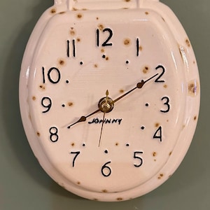 May include: A wall clock shaped like a toilet seat with a cream-colored ceramic finish and brown speckles. The clock face features black numbers from 1 to 12, gold-colored hands, and the word JOHNNY printed in black below the center hands.