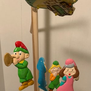 May include: A ceramic wind chime with a green and brown house at the top and four hanging figures. The figures are a green elf playing a trumpet, a blue fish, a green elf with a yellow hat, and a pink girl with a brown hat.