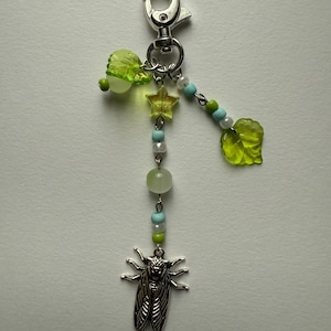 May include: A silver-toned keychain with a lobster clasp, adorned with green and blue beads, a yellow star, and a silver cicada charm. Features translucent green leaf and orb-shaped accents. A whimsical accessory.