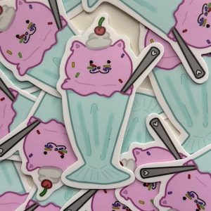 May include: A pile of stickers depicting a cartoon cat-shaped ice cream sundae. The pink ice cream has a cat face, sprinkles, and a cherry. The sundae is in a light blue glass with a spoon.