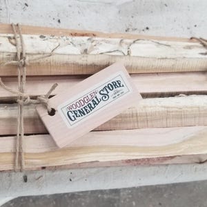 May include: A bundle of firewood tied with twine, featuring a wooden tag that reads "WOODGLEN GENERAL STORE". The wood has varying shades of brown and tan, with some pieces showing natural bark.