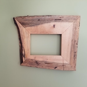 May include: Square wooden picture frame with a natural, rustic aesthetic. The frame is made from light-colored wood with dark, textured edges. The frame is mounted on a light green wall.
