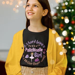 Op de afbeelding: Een persoon draagt een zwart t-shirt met de tekst "It's hard being a Polichinelle Girl in a Mother Ginger world" in paars en wit. De persoon draagt een gele cardigan en een beige rok. Op de achtergrond is een kerstboom te zien.