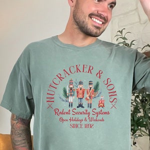 May include: Moss green t-shirt with a circular graphic featuring three nutcracker figures and the text "NUTCRACKER & SONS". The graphic also includes the text "Rodent Security Systems, Open Holidays & Weekends, SINCE 1892".