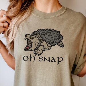 May include: Khaki t-shirt with a graphic of a snapping turtle and the text "oh snap".