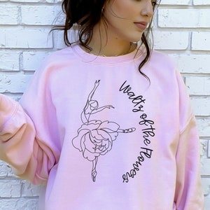 May include: Light pink sweatshirt with the text "Waltz of the Flowers" and a ballerina graphic. The sweatshirt is a solid light pink color. The graphic is a black line drawing of a ballerina and a flower.