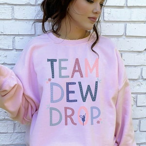 May include: A pink sweatshirt with the text "TEAM DEW DROP" in colorful letters with a small ballerina graphic. The letters are outlined with small dots.