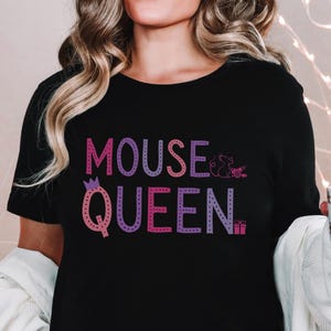 Op de afbeelding: Zwart T-shirt met de tekst "MOUSE QUEEN" in een roze en paarse gradiënt. Het ontwerp bevat een kroon, een muis en een geschenkdoos. Het shirt heeft een ronde hals.
