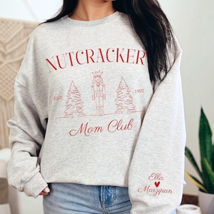 May include: Gray sweatshirt with a red graphic of a nutcracker, two Christmas trees, and the text "Nutcracker Mom Club Est 1892". The sleeve has the text "Ella Marzipan" with a red heart.