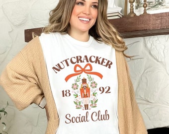 May include: A white t-shirt with a graphic that reads "Nutcracker Social Club 1892" in brown and orange. The design includes a nutcracker illustration and floral accents. The person is wearing a tan cardigan and light blue jeans.