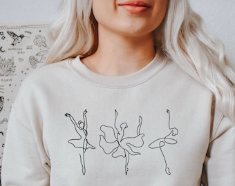 Op de afbeelding: Een beige sweatshirt met een zwarte lijntekening van drie ballerina's op een rij.