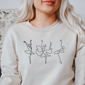 May include: A beige sweatshirt with a black line drawing of three ballerinas in a row.