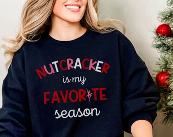 Peut inclure: Un sweat-shirt bleu marine avec le texte "NUTCRACKER is my FAVORITE season" en rouge et blanc. La personne porte un pantalon de pyjama à carreaux rouges et noirs. Un sapin de Noël avec des boules rouges est en arrière-plan.
