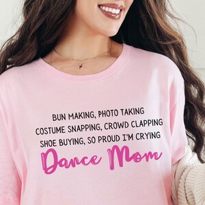 Op de afbeelding: Roze T-shirt met de tekst "BUN MAKING, PHOTO TAKING, COSTUME SNAPPING, CROWD CLAPPING, SHOE BUYING, SO PROUD I'M CRYING Dance Mom" in zwarte en roze letters.