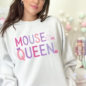 May include: White sweatshirt with the text "MOUSE QUEEN" in pink and purple, with a crown graphic on the letter "Q" and a mouse graphic. The sweatshirt is a crew neck style.