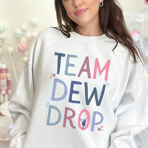 May include: A white sweatshirt with the text "TEAM DEW DROP" in blue, pink, and purple. The design includes a ballerina graphic and snowflake accents. The sweatshirt is a casual, comfortable style.
