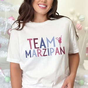May include: White t-shirt with the text "TEAM MARZIPAN" in pink, blue, and purple letters with white polka dots. A pink silhouette of a ballerina is next to the word "TEAM".