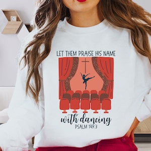 May include: White sweatshirt with a red and white illustration of a dancer on a stage with the text "Let them praise his name with dancing Psalm 149:3".
