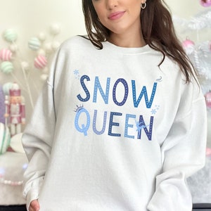 May include: White sweatshirt with the text "SNOW QUEEN" in blue and white lettering with snowflake and crown accents.