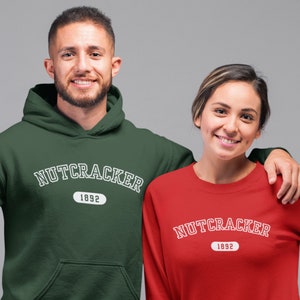 Op de afbeelding: Een persoon draagt een donkergroene hoodie en een ander een rode crewneck-trui. Beide sweatshirts hebben het woord "NUTCRACKER" in witte boogtekst boven het jaar "1892" in een witte ovaal.