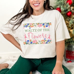 May include: Beige t-shirt with the text "Waltz of the Flowers" in green and pink, framed by a floral border. The shirt is paired with green sweatpants. The overall theme is ballet and floral design.