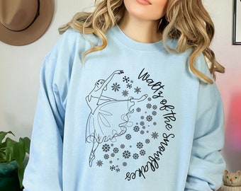 May include: Light blue sweatshirt featuring a black line drawing of a ballerina and snowflakes. The text "Waltz of the Snowflakes" is printed in black. The sweatshirt is a light blue color.