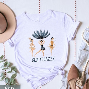May include: A white t-shirt with a graphic of three women in flapper dresses. The women are holding a large feather boa. The text "Keep it Jazzy" is printed below the graphic.