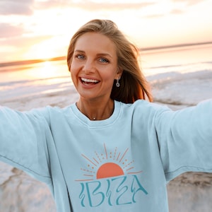 May include: A light blue sweatshirt featuring a graphic of a sunrise and the word "IBIZA" in a turquoise blue font.