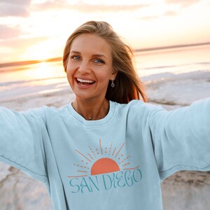 May include: A woman wearing a light blue sweatshirt with a graphic of a sunset and the text "SAN DIEGO" in teal blue.