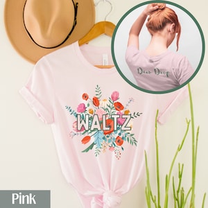 May include: A light pink t-shirt with the word "WALTZ" in a white and black font, surrounded by a colorful floral design. The back of the shirt has the words "Dew Drop" in a green font. A tan hat hangs in the background.