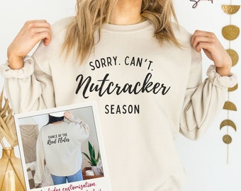 May include: A beige sweatshirt with the text "SORRY. CAN'T. Nutcracker SEASON" printed on the front. The back of the sweatshirt has the text "DANCE OF THE Reed Flutes" printed on it. The sweatshirt is being held by a person wearing blue jeans.